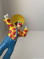 Side view of clown figurine showing detailed painted features, yellow shirt with red spots, blue pants, and black shoes with right arm pointing up.