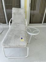 White metal cushioned patio chaise lounge with weathered cushion and small round glass-top side table.
