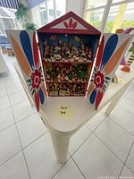 Front view with doors open showing three shelves of miniature figurines representing village scenes within the painted wooden house.