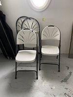 Photo of four folding chairs, two fully visible from front with white molded plastic seats and backs, two folded and leaning against wall behind. Chairs have black metal tubular frames and smooth plastic handles at the top back of chair backs. Floor is white painted concrete with some scuffs.