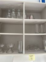 Glassware including clear glass bottles, short tumblers with playing card suits, tall tumblers, footed dessert cups, and glass plates arranged in a shelf compartment.