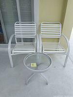 Two white metal outdoor chairs and one small round glass top table on a concrete floor outdoors.