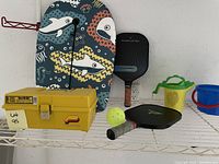 Photo of boogie board, two pickleball paddles, a yellow pump ball, tool box, and sand play buckets