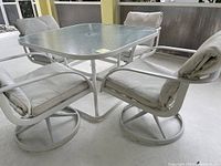 View of square textured glass tabletop with metal frame and four cushioned swivel chairs around it on patio floor.