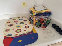 A corner countertop pictured with a pile of colorful cloth napkins, bright patterned placemats, and several sets of napkin rings: glass, ceramic bird-shaped, and dark blue ceramic.