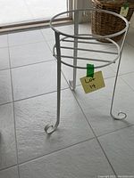 Photo shows a white metal plant stand with a circular holder and three curled legs on tiled floor.
