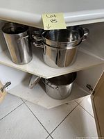 Polished stainless steel pots and pans on a corner shelf, including a tall cylindrical pot, a large pot with a steamer insert, and medium pots below.