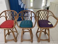 Front view of three rattan bar stools with colored cushions in red, teal, and purple showing the lattice design and armrests.