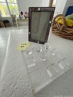 Photo shows the glass framed Tic Tac Toe game board with stained glass border and the transparent acrylic playing pieces arranged in front on a white textured surface.