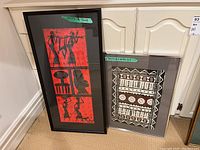 Two vertical framed art pieces adjacent on floor against cabinet. Left piece black frame with red and black figure art signed. Right piece silver frame with black and white patterned art.