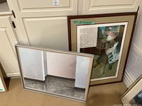 Two framed prints leaning against a white cabinet. Larger piece with wooden frame scratched, smaller with silver frame.