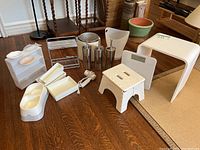Photo showing white plastic shower stool with wooden legs, folding step stool, trash bin, soap dishes, metal toilet scrubbers, white pet dish, and baskets on wooden floor near furniture.