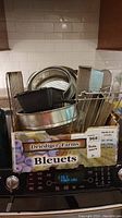 Photo shows a box labeled 'Bleuets' holding various metal bakeware pieces including cake pans, loaf pans, baking trays and a wire cooling rack.