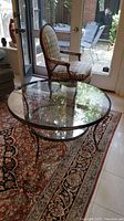 Full view of round glass coffee table with metal base and claw feet on a patterned rug near a checkered upholstered armchair.