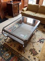 Angle side view of metal frame coffee table with glass insert sitting on a patterned rug in a living room. Sofa and cabinet visible in background.