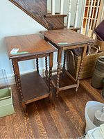 Photo showing two wooden end tables positioned side by side near a staircase, one with twisted spindle legs and the other with straight carved legs, on a hardwood floor.