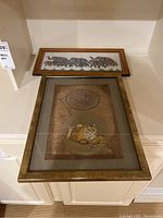 Photo of two framed artworks; top frame contains fabric art with stylized animals, bottom frame contains painted fabric art with tiger and script.
