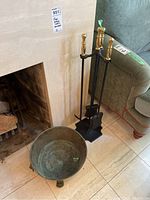 Photo showing the three-piece fireplace tool set with brass handles on a black iron stand next to a metal footed bucket on the floor by a fireplace.