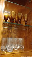 Amber-colored wine glasses on upper shelf and clear glass tumblers on lower shelf inside wooden cabinet