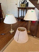 Two table lamps and one spare lampshade placed on floor. One lamp with round polished metal base and pleated fabric shade, one lamp with rectangular metal and glass base and fabric shade.