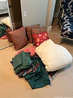 Photo of five decorative pillows in various patterns and colors, a white quilted blanket, a pink fringed blanket, and a stack of dark green and patterned bed linens on a beige carpet next to a mirror and a chair.