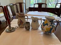 Photo showing three pitchers, a small pottery cup, and a metal candle holder with red candle displayed on a table with chairs and window background.