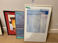 Three framed posters leaning against a wall: San Francisco Museum of Modern Art abstract poster, Van Gogh print, and Mary Pratt poster.
