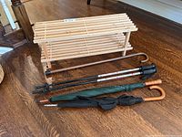 Wooden two-tier slatted shoe rack with walking poles, two umbrellas, and wooden cane placed on hardwood floor.