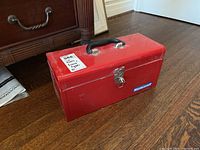 Closed red metal Mastercraft toolbox on wooden floor with handle and latch visible