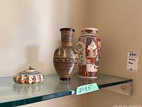 Photo of two ceramic vases and one round lidded ceramic box on a glass shelf.