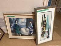Two framed watercolours leaning against wall, one portrait orientation with bottles, one landscape with women and dog