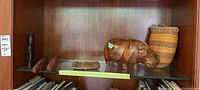 Overall view of wood art pieces and basket on shelf, includes carved hippo, wooden claves, carved head, ceramic dish, and woven basket