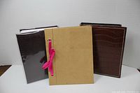 Three small photo albums, two dark brown textured and one tan suede with pink ribbon, shown upright and closed.