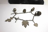 Charm bracelet laid flat showing entire chain and all six unique charms including heart, leaf shapes, cross, teardrop bead and cage charm.