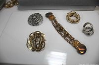 Five scarf pins/clips and a chain sweater holder displayed on a white surface. Visible details include metal tones, decorative elements, and overall condition.