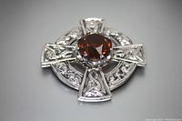 Clear close-up of the front of a silver-tone Celtic cross skirt pin or brooch featuring a faceted topaz colored central stone and intricate metal design.