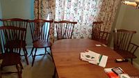 Full view of six maple wood dining chairs around a wooden table with floral curtains in background.