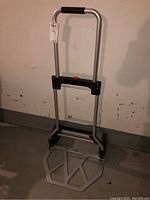 Folding dolly fully extended, metal frame and base with black plastic parts, foldable handle with foam grip visible.