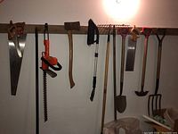 Tools hanging on wooden rack showing saws, hedge trimmer, axe, broom, rake, pitch fork, hoe, and shovel in a garage setting.