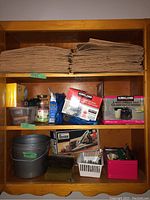 Wide view of wooden shelf displaying stacks of brown yard waste bags, Kirkland brand garbage bags boxes, plastic buckets, and containers with miscellaneous tools and items.