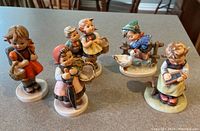 Five Hummels porcelain figurines displayed on a tabletop, showing various children in different poses and activities.