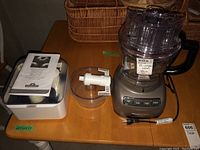 Full view of KitchenAid food processor with bowl, lid, and white attachments on wooden table with user manual visible under one attachment.