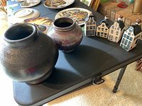 Photo showing two ceramic vases at left side of table, one larger with dark glaze and one smaller with reddish patina, and four KLM Delft Blue ceramic house-shaped bottles at right side.