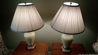 Two ceramic lamps with cream bases and pleated off-white fabric shades side by side on two wooden tables.