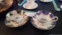 Two Paragon teacups and saucers displayed side by side on a black tablecloth, showing black/gold/rose and blue/gold floral designs.