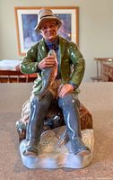 Front view of Royal Doulton A Good Catch figurine, showing fisherman holding fish seated on log.