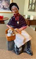Front view of the Royal Doulton figurine showing the woman seated on a bench, wearing a dark bonnet and shawl, holding a basket with a white dog.