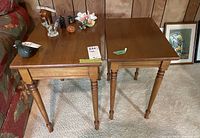 Two wooden nested side tables with turned legs placed side by side against a wood panel wall and carpeted floor, one larger and one smaller.