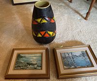 Photo showing the dark ceramic vase with vibrant orange, yellow, and red geometric triangle patterns next to two framed Keirstead nature prints, all resting on beige carpet.