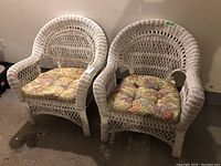 Two white painted wicker armchairs with floral patterned fabric cushions shown side by side from front angle.